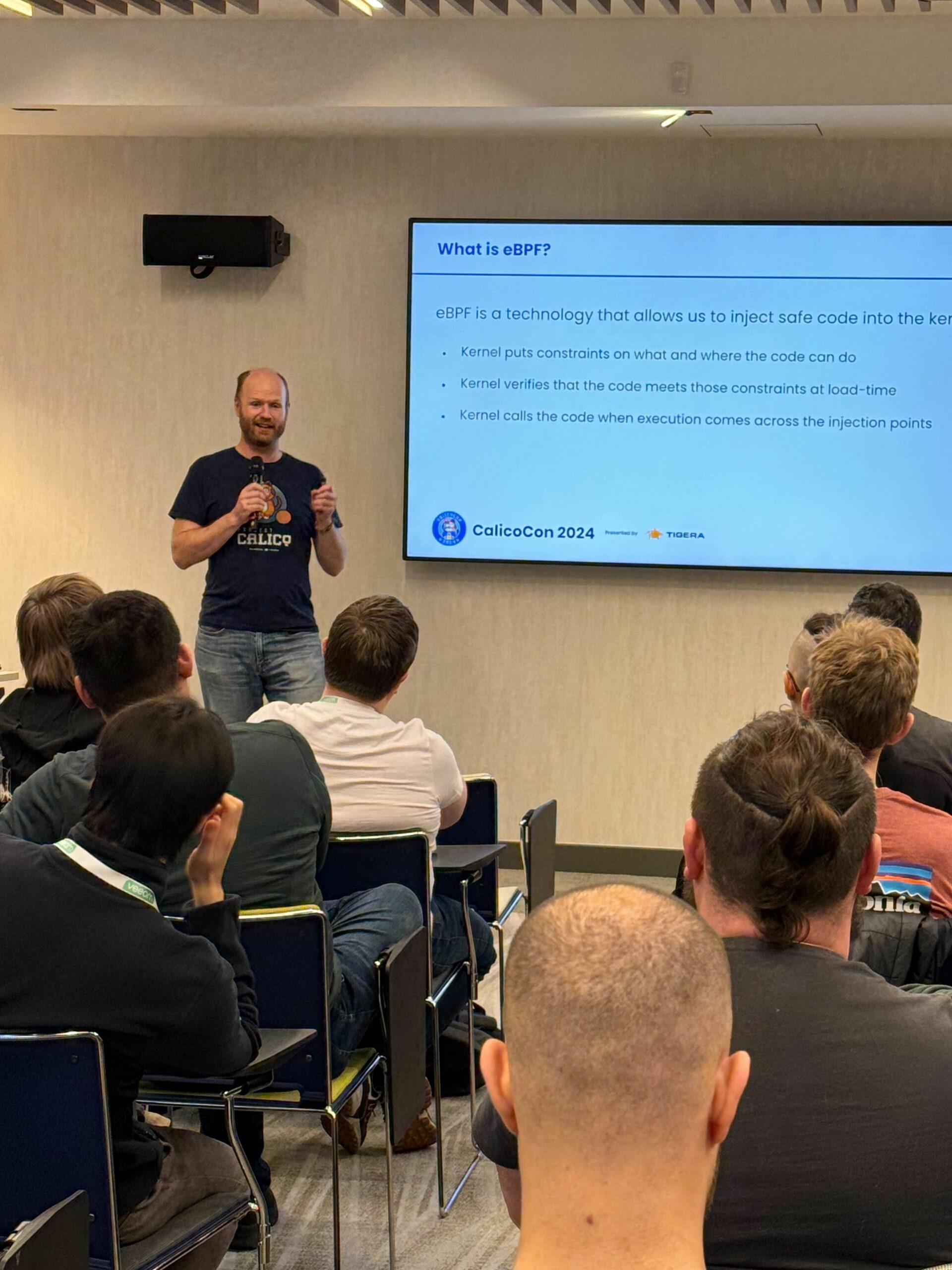 A speaker presents at CalicoCon 2024, explaining eBPF technology to an audience seated in chairs. A screen displays the presentation title and bullet points