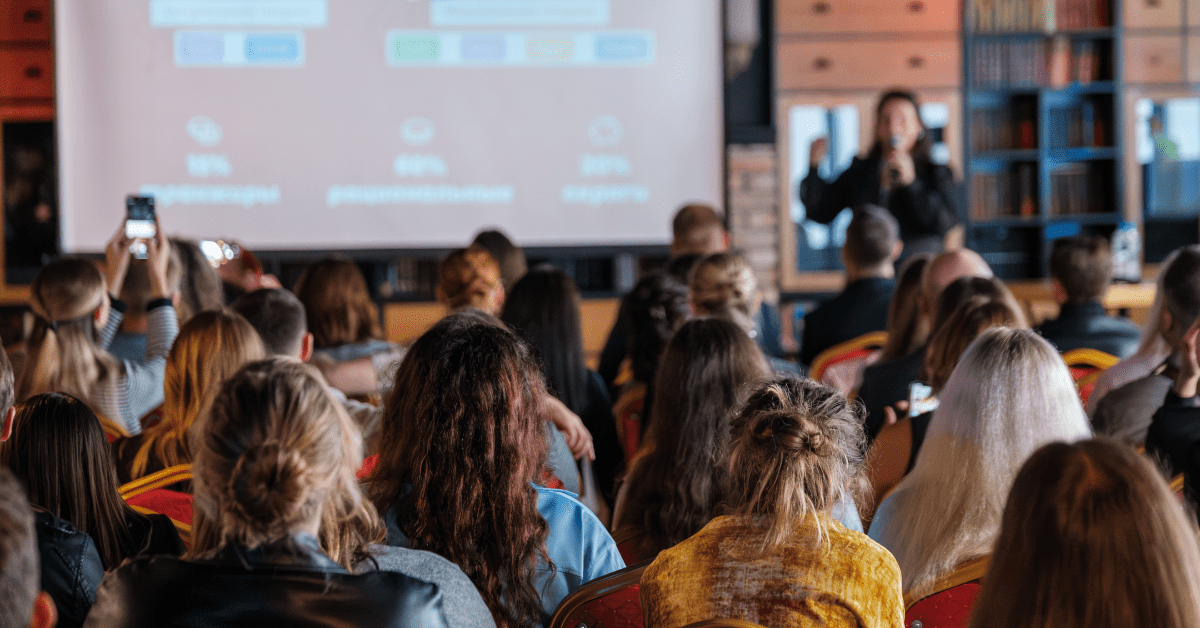 An audience watches a speaker present slides on a large screen.