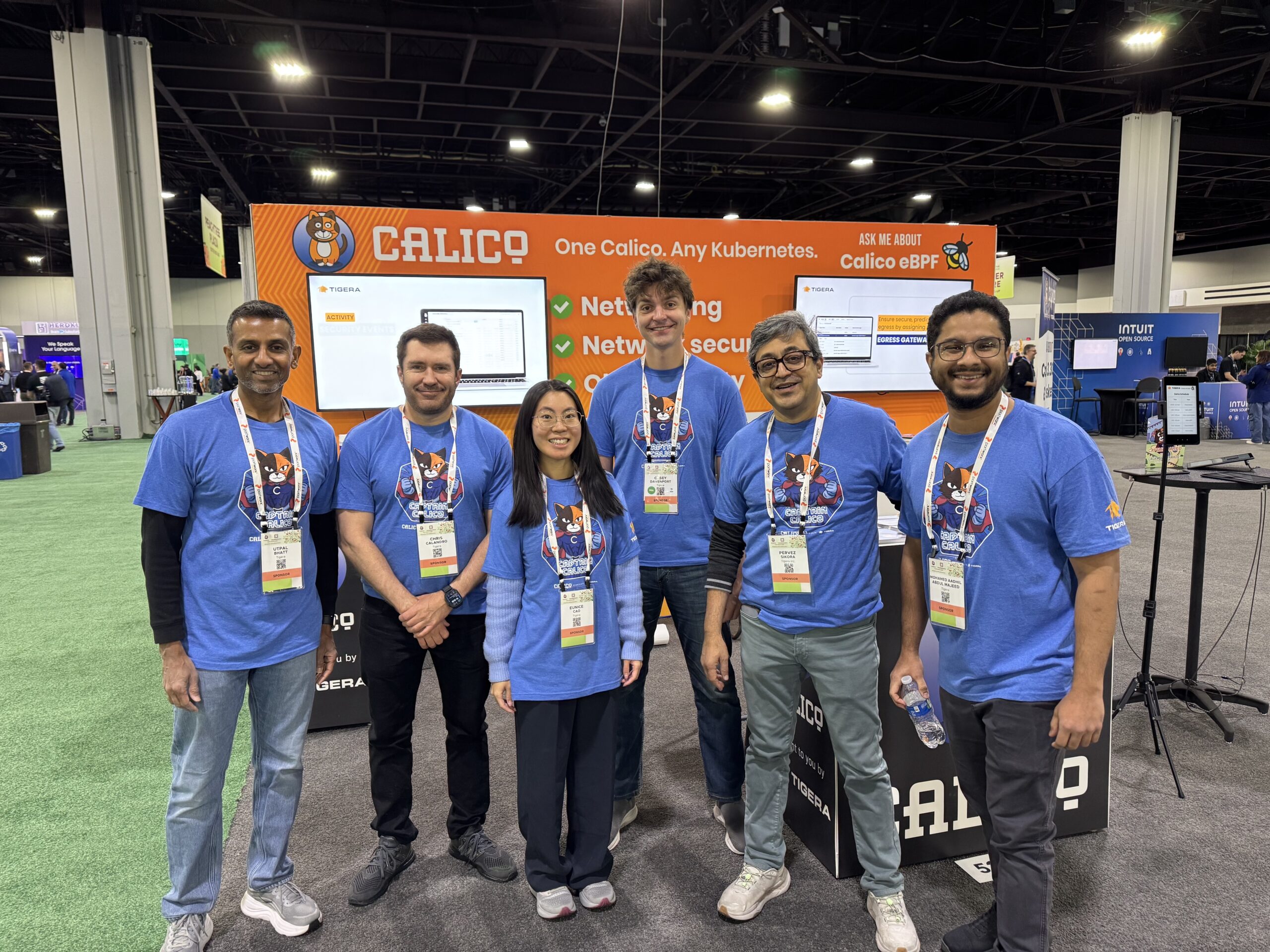 Six smiling Calico team members pose at their orange KubeCon booth.